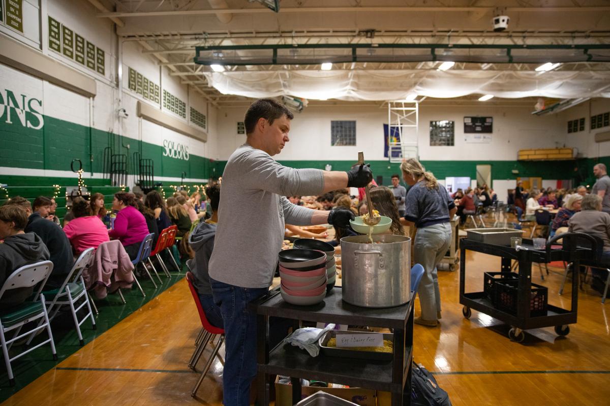 A teacher ladles soup into a bowl. In the background, many students are seated at long tables in a school gymnasium.