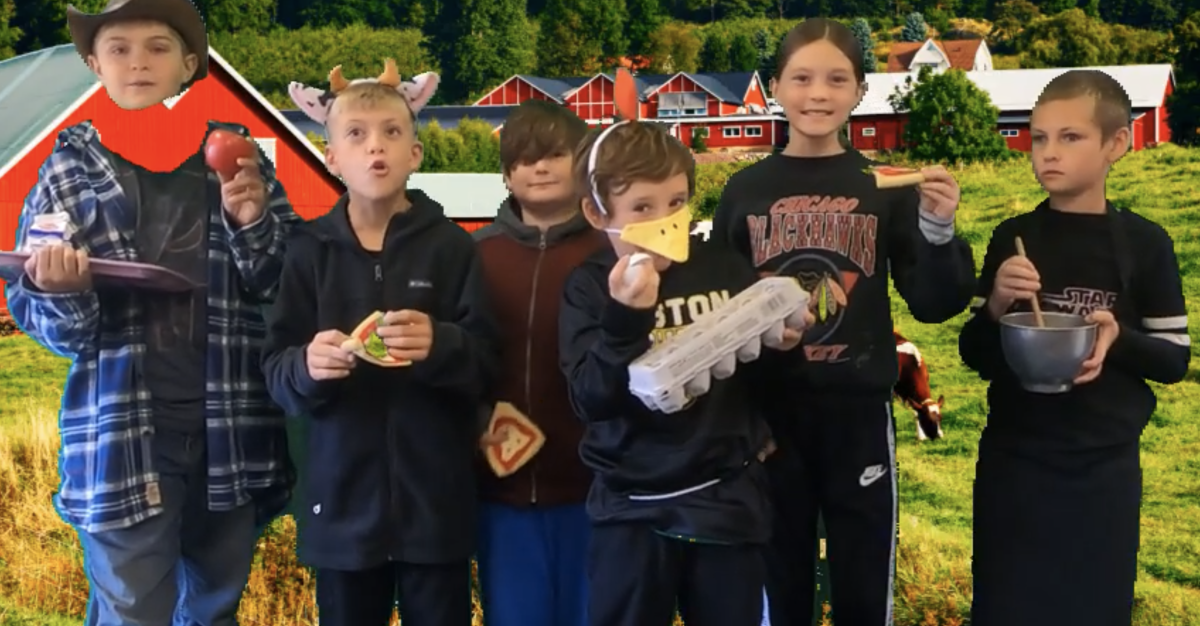 A group of students stands in front of a backdrop featuring a farm scene with red barns. They are holding various food items and wear playful accessories like a hat and animal ears.