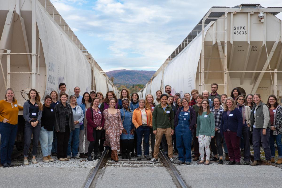 Group of people standing between two rows of white train cars on railroad tracks, with a scenic mountain backdrop