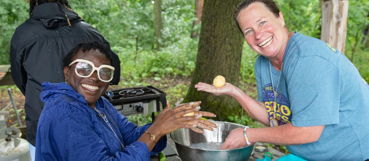 Two educators sit outdoors at a wooden table making wheat rolls