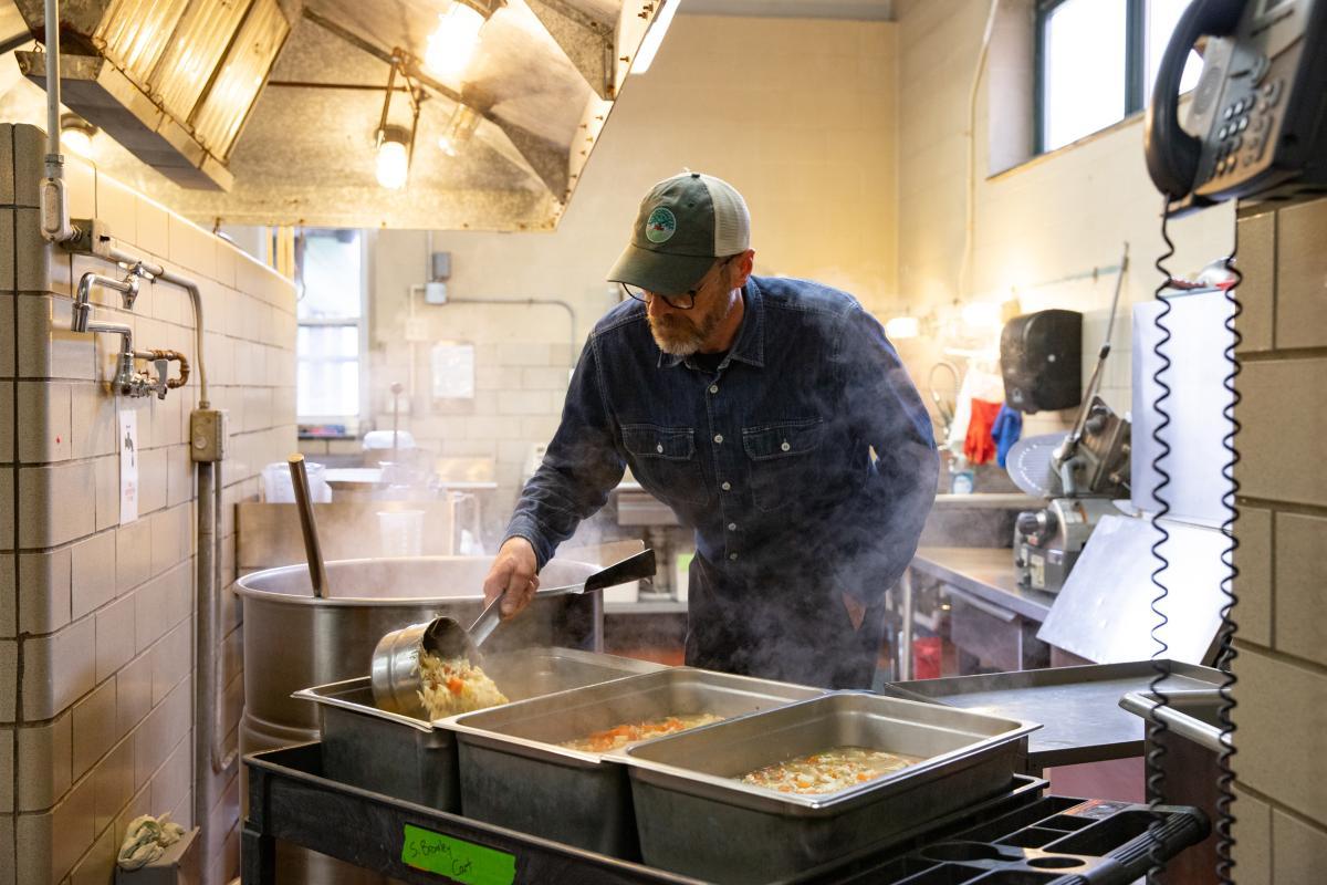 A man ladles soup into large hotel pans in a school kitchen. A man ladles soup into large hotel pans in a school kitchen.