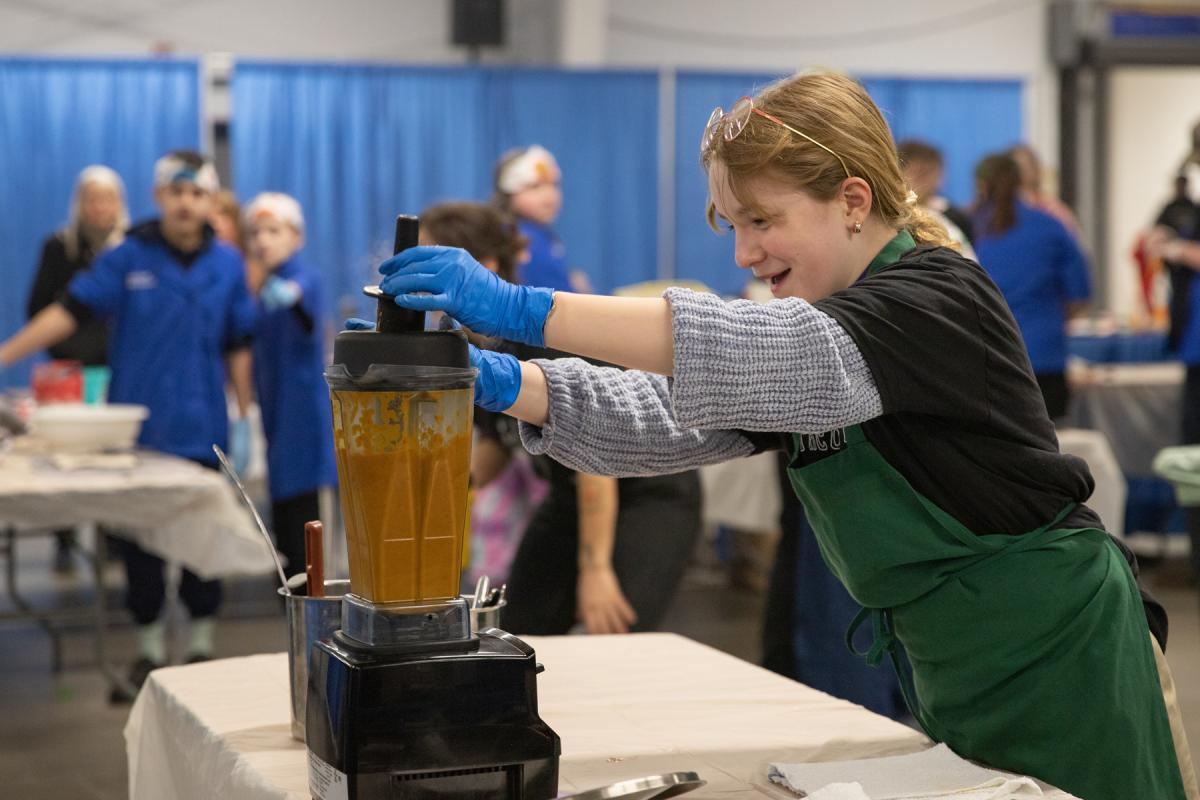 a young person smiles and holds the top of a blender while pureeing an orange liquid. a young person smiles and holds the top of a blender while pureeing an orange liquid.