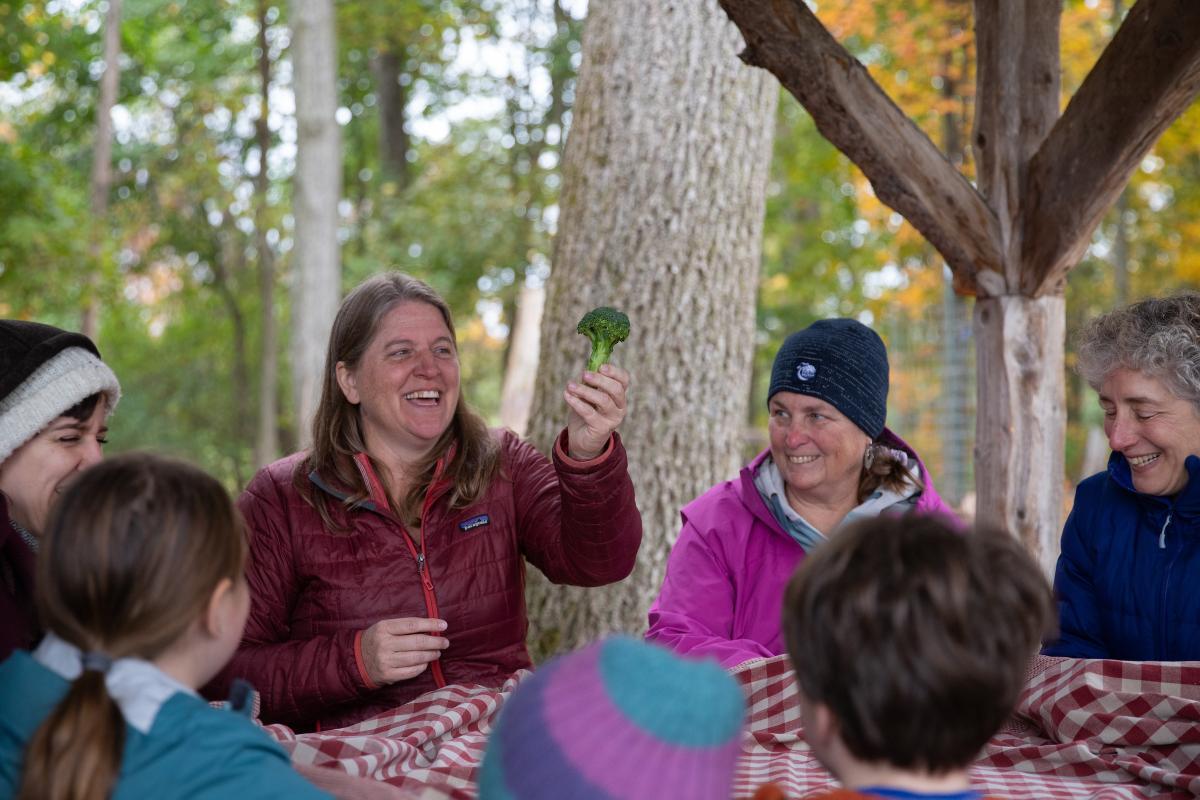 A woman holds up a piece of broccoli, she is part of a group seated in at a picnic table A woman holds up a piece of broccoli, she is part of a group seated in at a picnic table