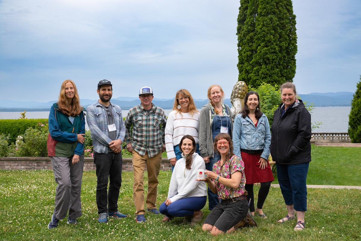 A group of people is gathered in a garden setting with a lake and mountains in the background A group of people is gathered in a garden setting with a lake and mountains in the background