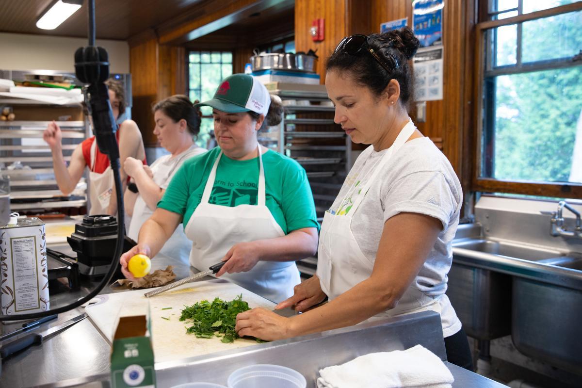 Three people wearing aprons prepare food in a commercial kitchen, chopping herbs and slicing lemons. Three people wearing aprons prepare food in a commercial kitchen, chopping herbs and slicing lemons.
