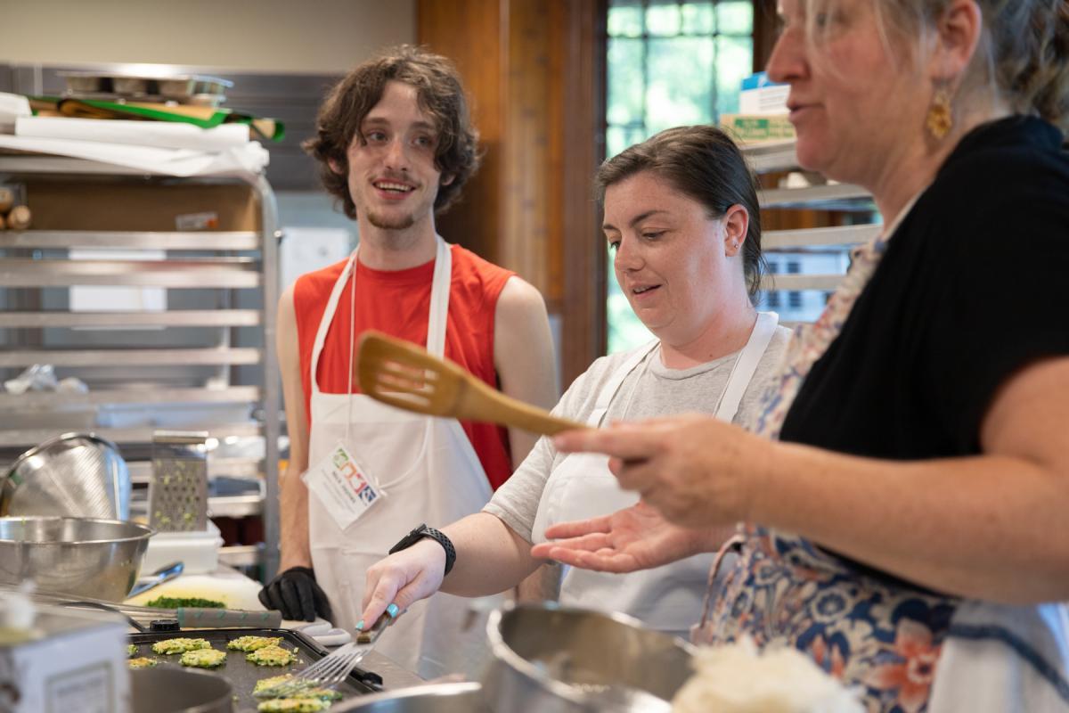 Three participants are engaging in a cooking class, preparing food together. One individual handles a spatula over a stovetop, while another holds a wooden spoon, and a third observes their actions closely.