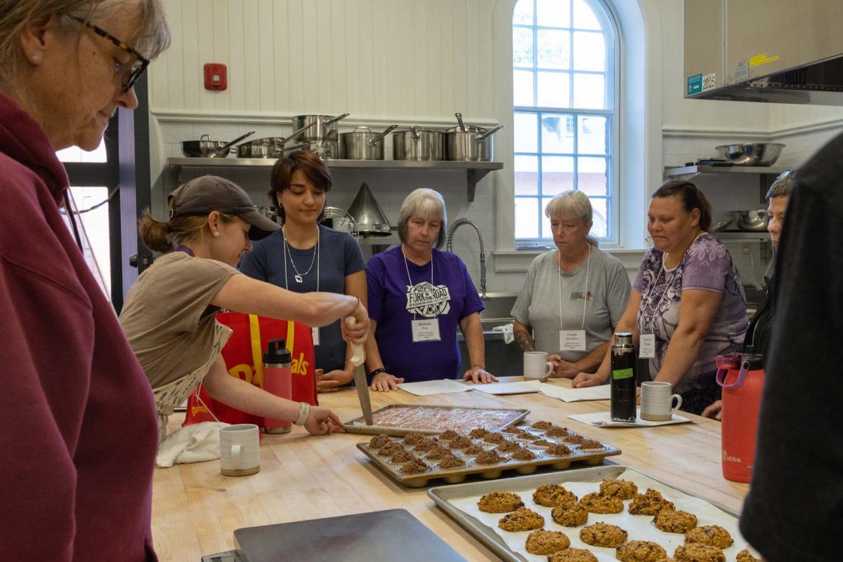 A group of people standing around a commercial kitchen table, watching as one person demonstrates with a knife. The table has trays of baked goods, including cookies. A group of people standing around a commercial kitchen table, watching as one person demonstrates with a knife. The table has trays of baked goods, including cookies.
