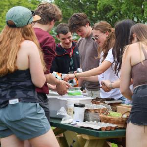 A group of young people gathers around a picnic table outdoors, engaged in planting seedlings. A group of young people gathers around a picnic table outdoors, engaged in planting seedlings.