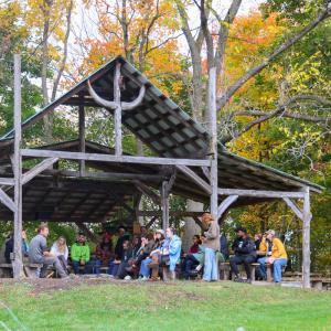 A group of people seated under a rustic open wooden shelter in a garden setting. A group of people seated under a rustic open wooden shelter in a garden setting.
