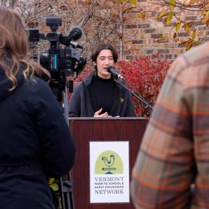 A student is speaking at an outdoor event, standing at a podium with a microphone. A student is speaking at an outdoor event, standing at a podium with a microphone.