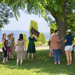 A group of people stand on grass under a large tree by a lake, with some holding and gesturing towards a "Community" sign. A group of people stand on grass under a large tree by a lake, with some holding and gesturing towards a "Community" sign.