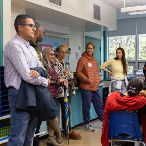 A group of people stand in a classroom, engaging with three seated students. A group of people stand in a classroom, engaging with three seated students.