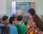 Three children and one adult at a milk dispensing station with a sign reading "Fresh, Organic Vermont Milk supplied by Miller Farm in Vernon, VT."