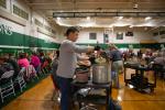 A teacher ladles soup into a bowl. In the background, many students are seated at long tables in a school gymnasium.