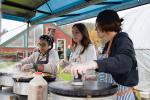 Three people cooking at an outdoor stall with a farm visible in the background.