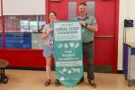 Two individuals holding a banner reading "Local Food Champion 2024-2025" inside a school cafeteria. The banner highlights the Essex Westford School District's support for local Vermont farms.