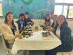 Group of six students smiling at a lunch table in a brightly-lit room with a playful underwater-themed mural in the background. They are enjoying various meals neatly arranged on the table.