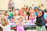 Group of children enjoying lunch together at school cafeteria tables, some looking at the camera and smiling.