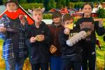 A group of students stands in front of a backdrop featuring a farm scene with red barns. They are holding various food items and wear playful accessories like a hat and animal ears.
